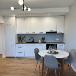 HOME INVEST PARTNERS SPÓŁKA Z OGRANICZONĄ ODPOWIEDZIALNOŚCIĄ - Jasna kuchnia z białymi szafkami, drewnianym blatem, szarą ścianą nad blatem i okrągłym stołem z szarymi krzesłami, oświetlona nowoczesną lampą z kulistymi kloszami.