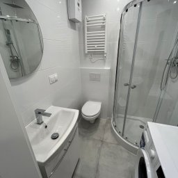 HOME INVEST PARTNERS SPÓŁKA Z OGRANICZONĄ ODPOWIEDZIALNOŚCIĄ - Wnętrze nowocześnie urządzonej łazienki z kabiną prysznicową, umywalką z szafką, toaletą, grzejnikiem drabinkowym i pralką, utrzymane w bieli i szarości.