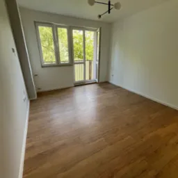 Puste, jasne pomieszczenie z drewnianą podłogą, białymi ścianami, oknem balkonowym z widokiem na zieleń i nowoczesną, czarną lampą sufitową.