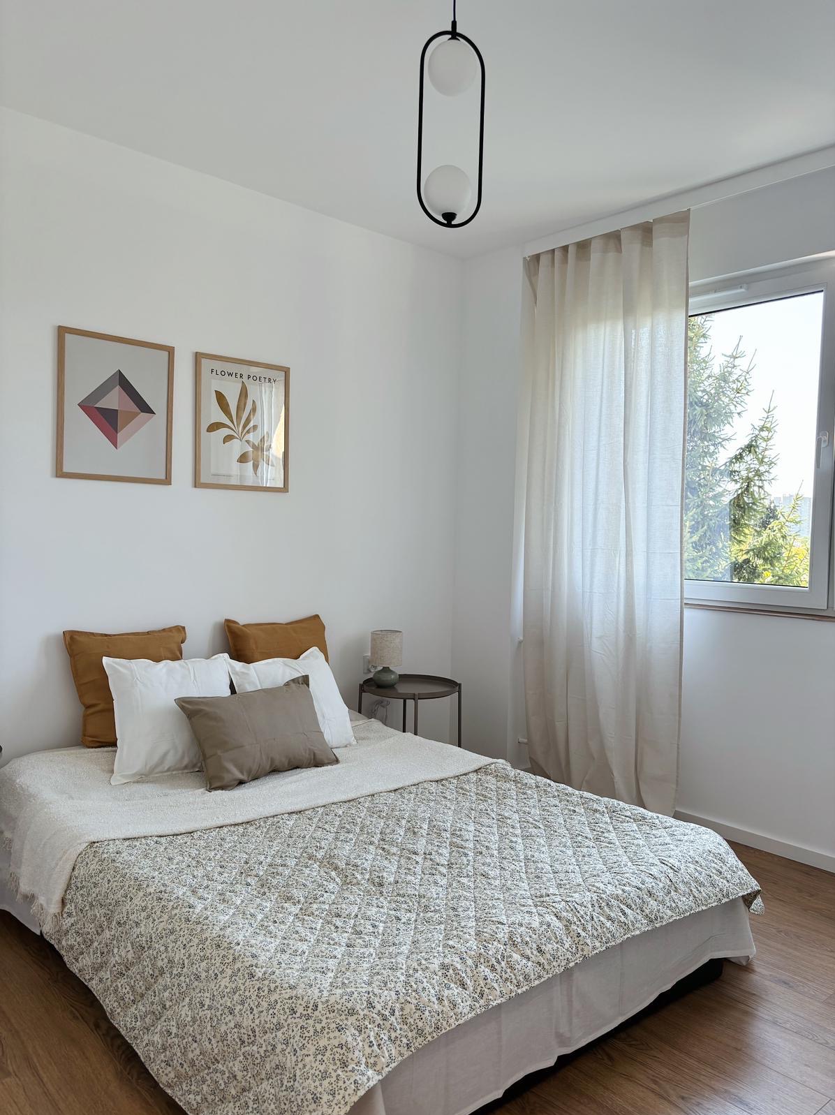 Jasna sypialnia z łóżkiem, wzorzystą narzutą, poduszkami w odcieniach brązu i bieli, stolikiem nocnym i widokiem na zieleń za oknem.