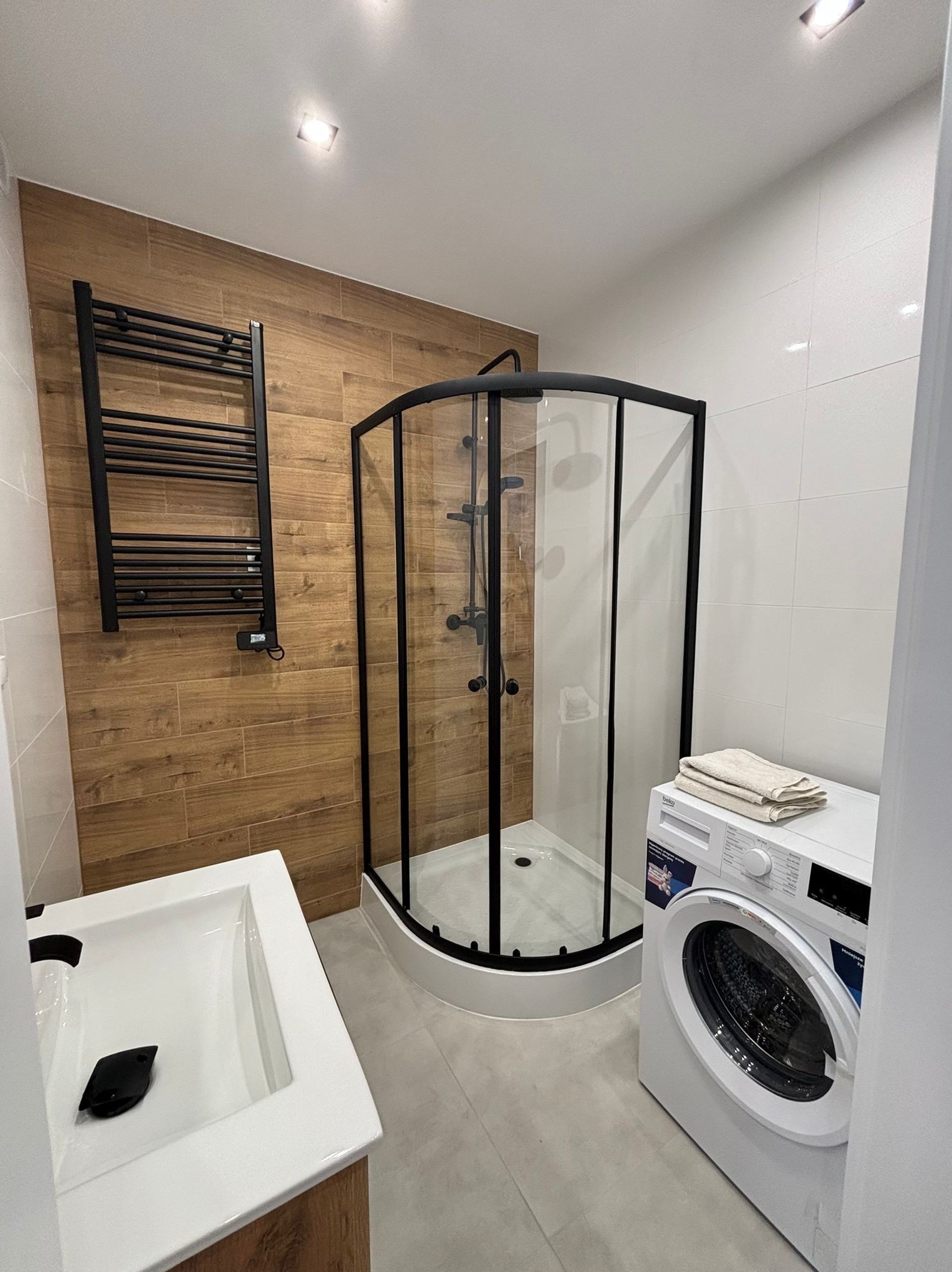 Nowoczesna łazienka z kabiną prysznicową narożną w czarnej ramie, grzejnikiem drabinkowym na brązowej ścianie imitującej drewno, białą umywalką i pralką z ręcznikami na górze.