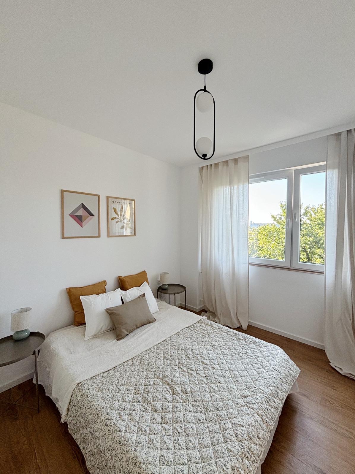 Jasna sypialnia z łóżkiem z pikowaną narzutą w drobne kwiatki, drewnianą podłogą i minimalistyczną lampą wiszącą o owalnym kształcie.