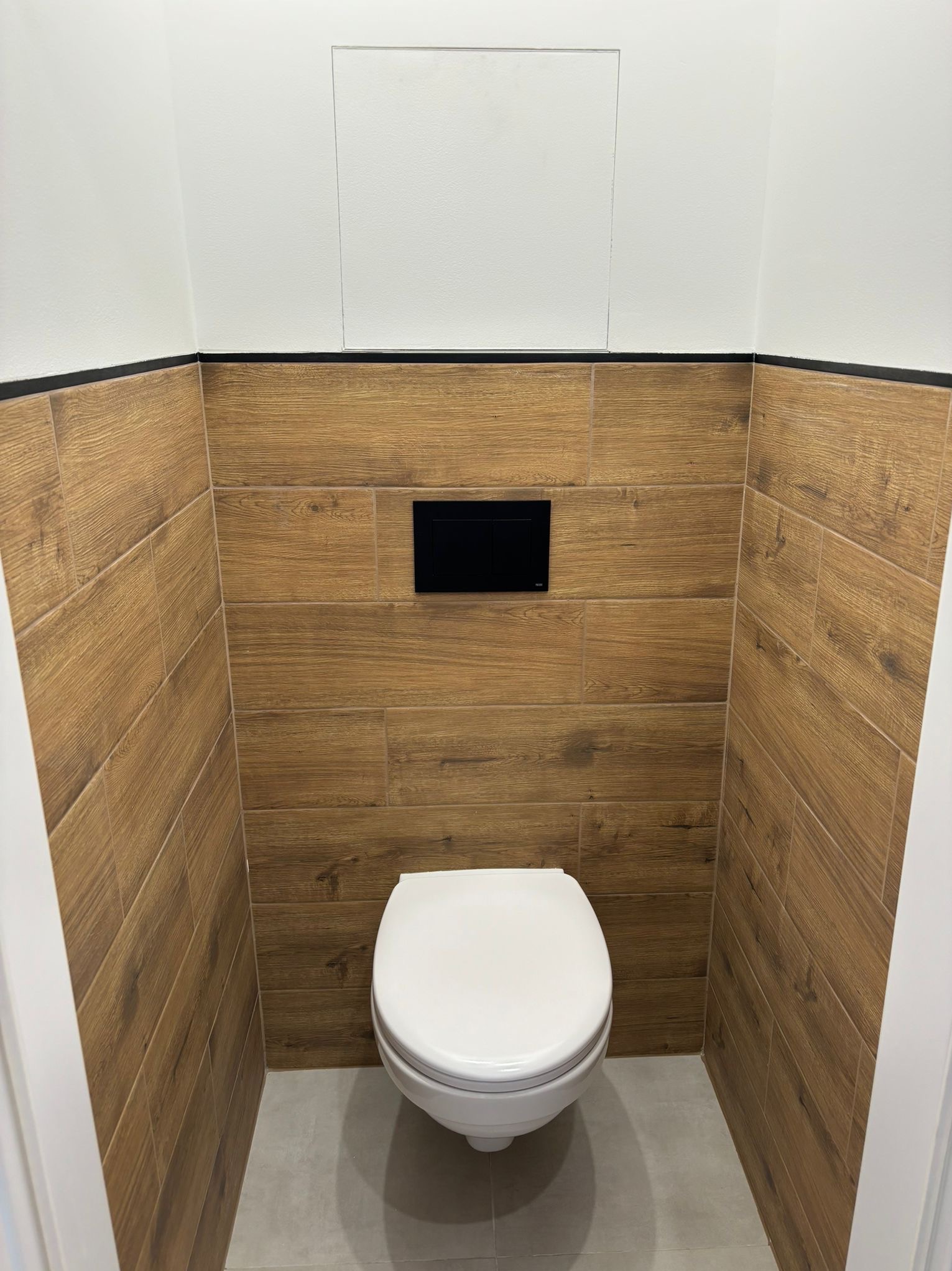 Nowoczesna toaleta z drewnopodobnymi płytkami ściennymi ułożonymi poziomo, czarny panel sterujący spłuczką, biała ceramika sanitarna i szara podłoga.