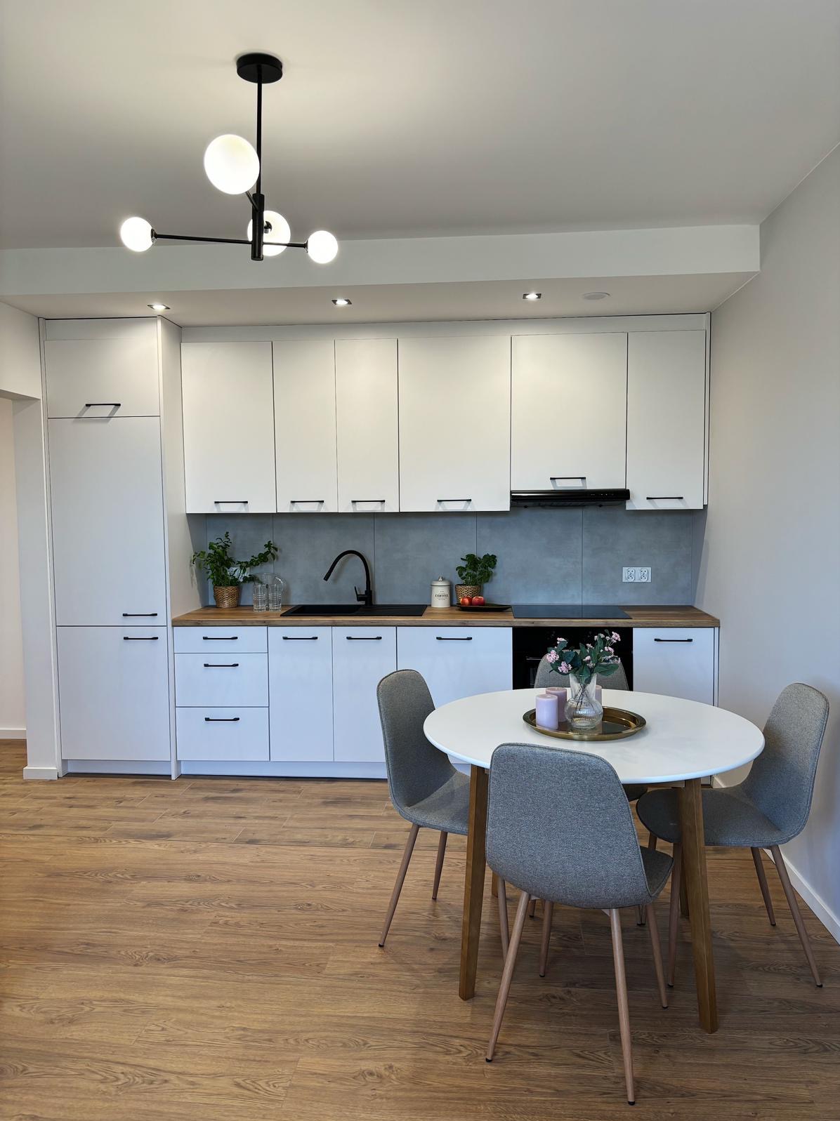 Jasna kuchnia z białymi szafkami, drewnianym blatem, szarą ścianą nad blatem i okrągłym stołem z szarymi krzesłami, oświetlona nowoczesną lampą z kulistymi kloszami.