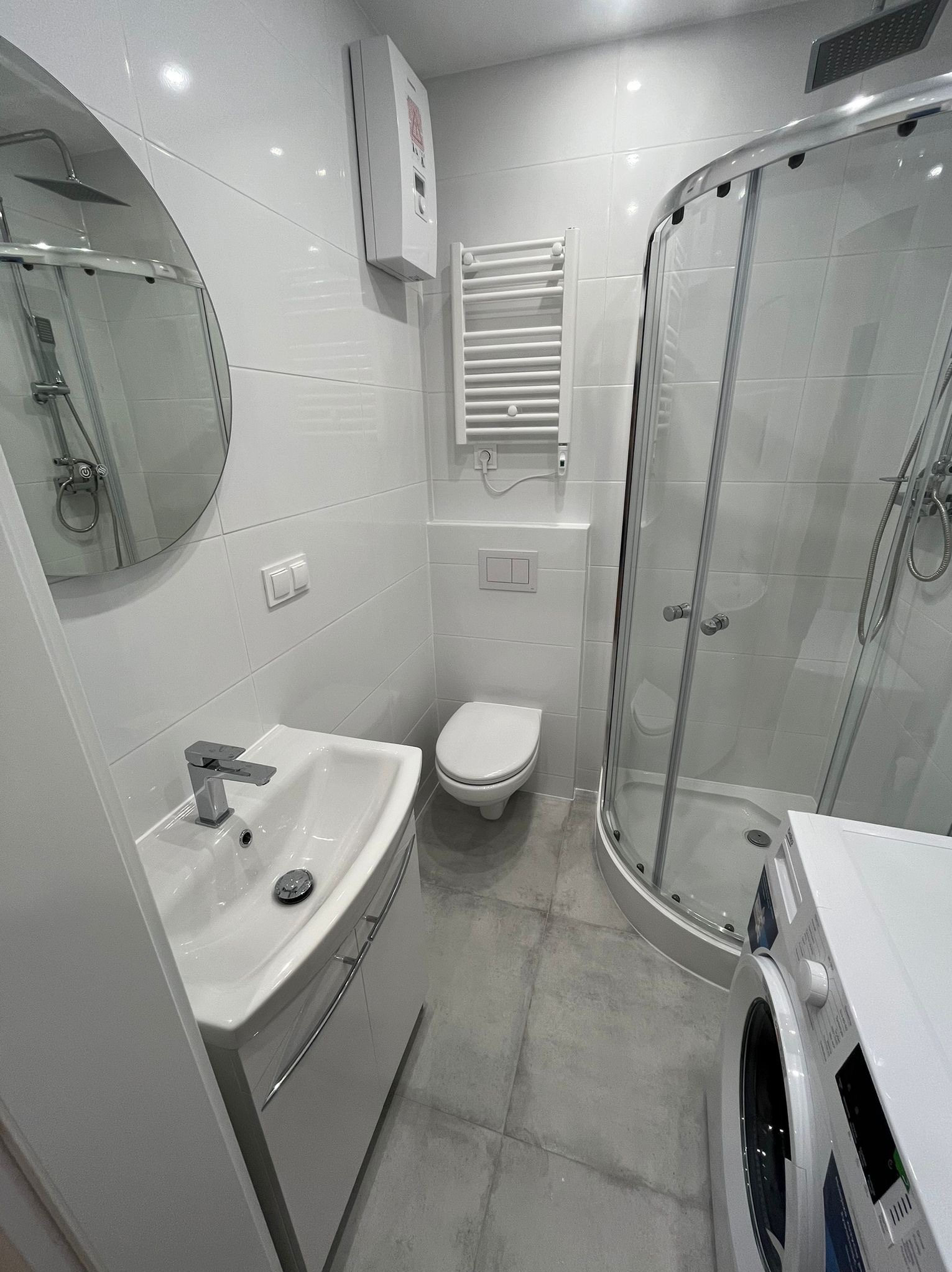 Wnętrze nowocześnie urządzonej łazienki z kabiną prysznicową, umywalką z szafką, toaletą, grzejnikiem drabinkowym i pralką, utrzymane w bieli i szarości.