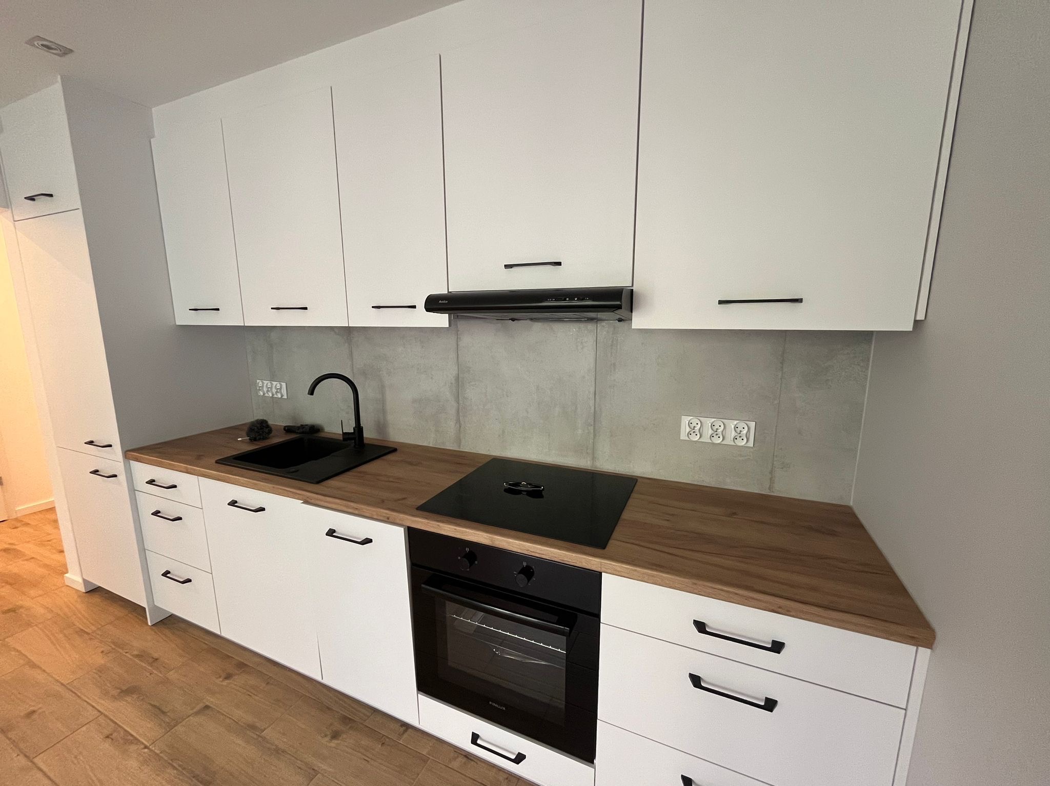 Nowoczesna, jasna kuchnia z białymi szafkami, drewnianym blatem, czarną armaturą i płytą indukcyjną, wykończona szarymi płytkami imitującymi beton.
