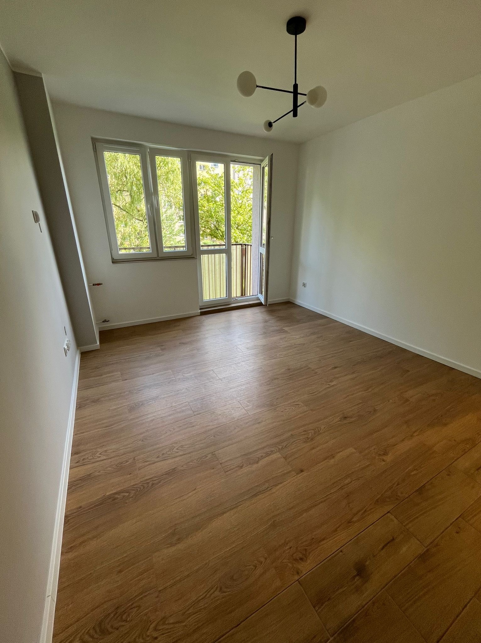 Puste, jasne pomieszczenie z drewnianą podłogą, białymi ścianami, oknem balkonowym z widokiem na zieleń i nowoczesną, czarną lampą sufitową.