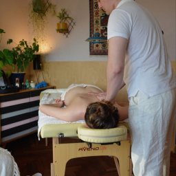 Jarosław Nemno - Osoba w białym ubraniu wykonuje masaż pleców pacjentki leżącej na profesjonalnym stole do masażu marki Aveno Life w jasnym pomieszczeniu z roślinami doniczkowymi i dekoracyjnym kilimem na ścianie.