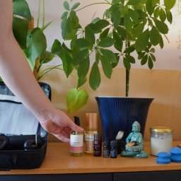 Jarosław Nemno - Zestaw do automasażu z pistoletem, olejkami i bańkami próżniowymi, ustawiony na drewnianej komodzie, z zielonymi roślinami w tle i figurką Buddy.