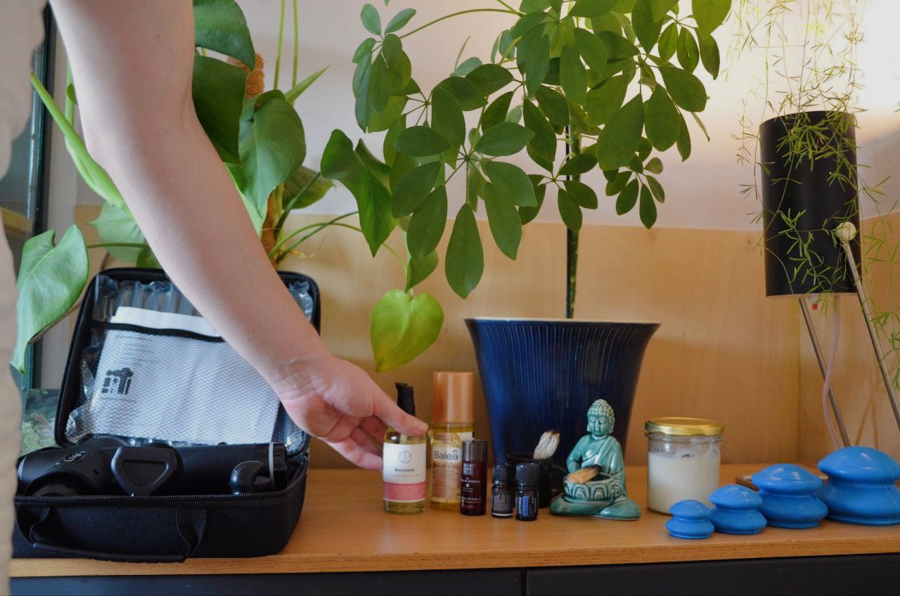 Zestaw do automasażu z pistoletem, olejkami i bańkami próżniowymi, ustawiony na drewnianej komodzie, z zielonymi roślinami w tle i figurką Buddy.