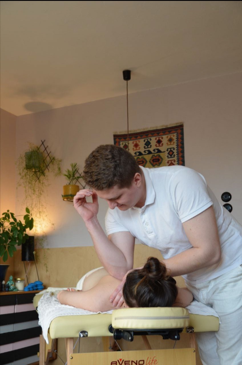 Mężczyzna w białym uniformie wykonuje masaż łokciem na plecach kobiety leżącej na profesjonalnym stole do masażu w gabinecie z roślinami doniczkowymi i wzorzystym dywanikiem na ścianie.