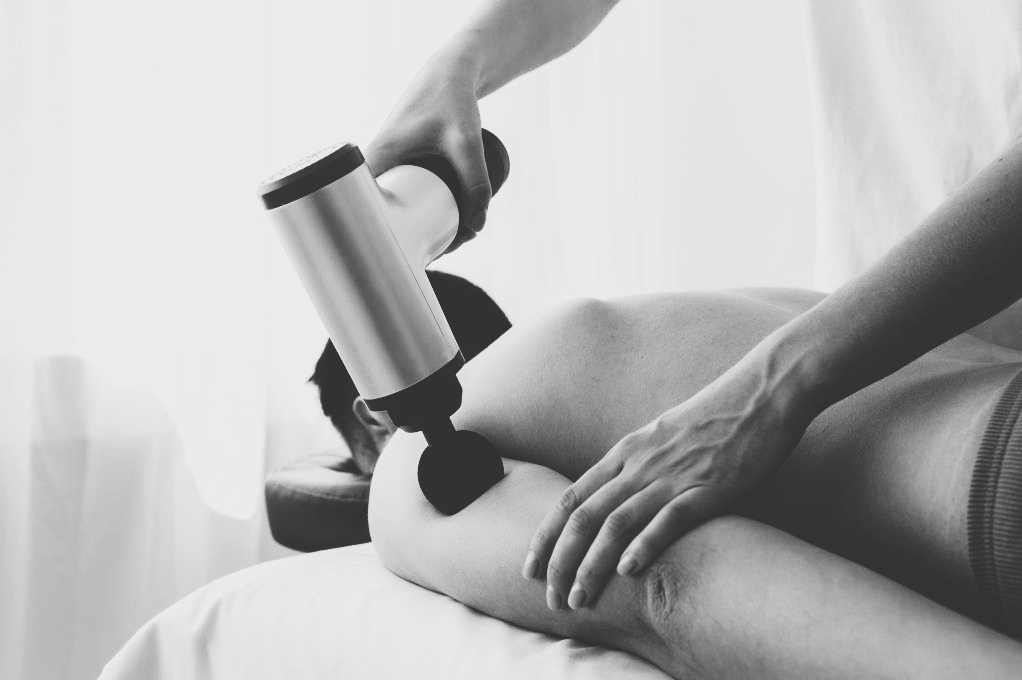 Czarno-białe zdjęcie przedstawia masażer wibracyjny używany na ramieniu pacjenta leżącego na białym prześcieradle, dłoń terapeuty stabilizuje urządzenie.