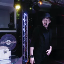 Iluzjonista w czarnym stroju i kapeluszu, z mikrofonem nagłownym, stoi na scenie z dekoracjami klubowymi i oświetleniem.