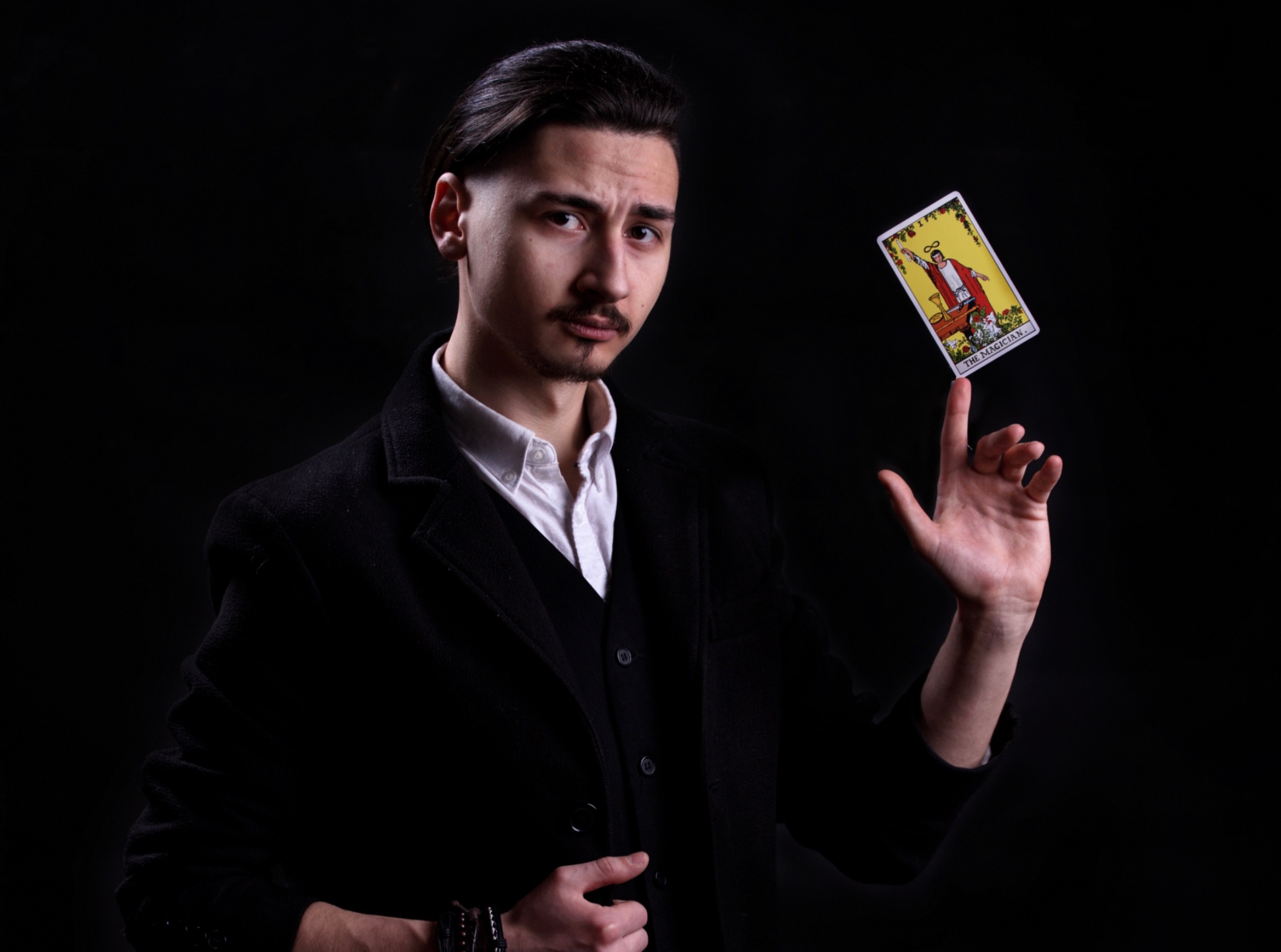 Młody mężczyzna w czarnym płaszczu balansuje kartą tarota 'Magik' na palcu, patrząc wprost w obiektyw na czarnym tle.