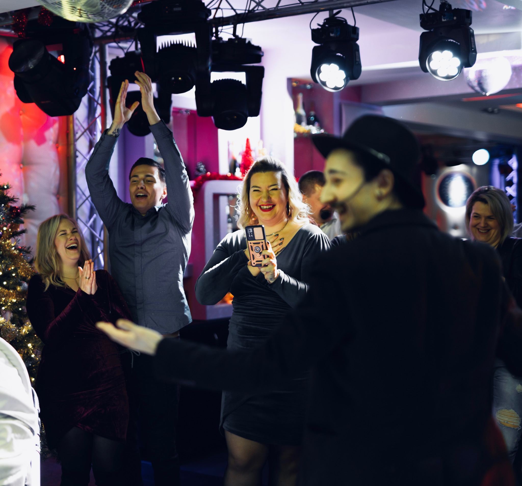 Iluzjonista w czarnym garniturze i kapeluszu występuje przed rozbawioną publicznością podczas imprezy, oświetlony reflektorami.