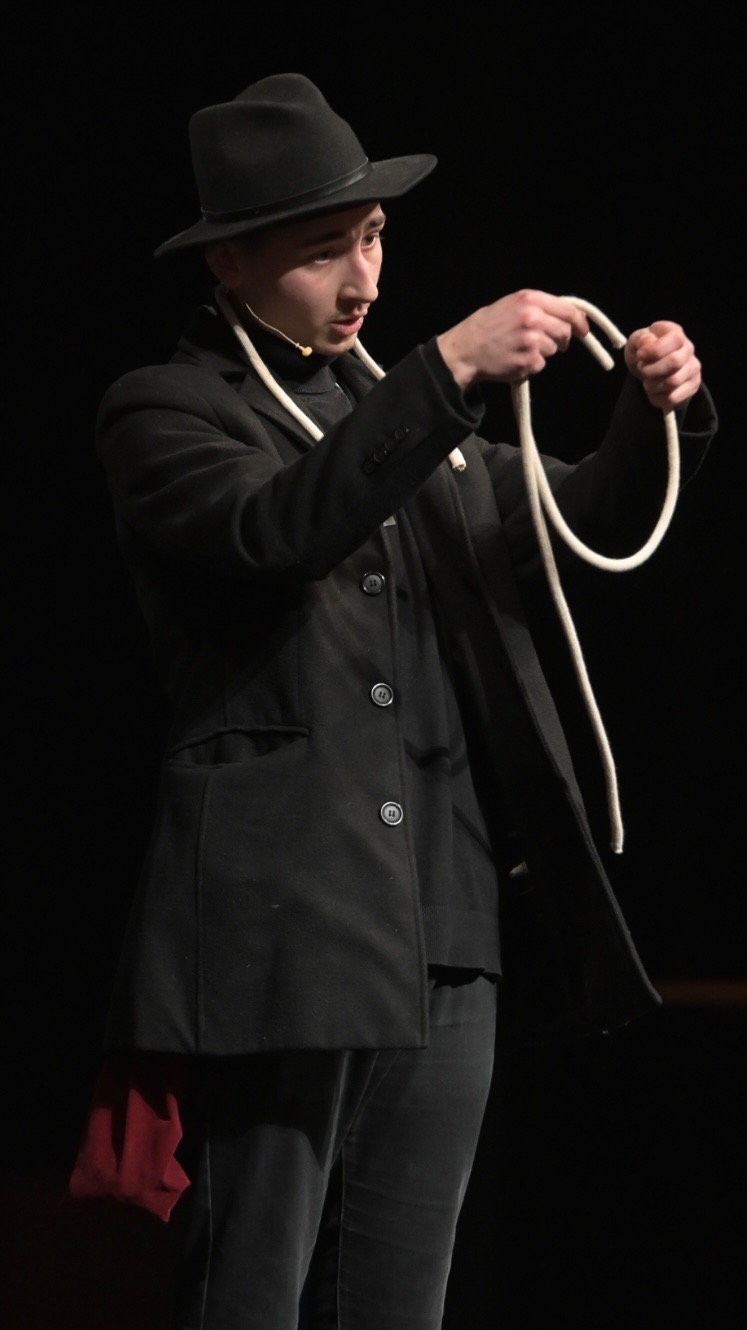 Młody iluzjonista w czarnym kapeluszu i płaszczu, prezentujący trik z białym sznurem na scenie z ciemnym tłem, z czerwonym rekwizytem wystającym z kieszeni.