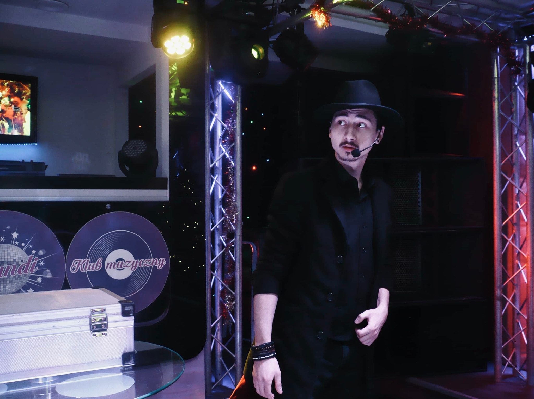 Iluzjonista w czarnym stroju i kapeluszu, z mikrofonem nagłownym, stoi na scenie z dekoracjami klubowymi i oświetleniem.