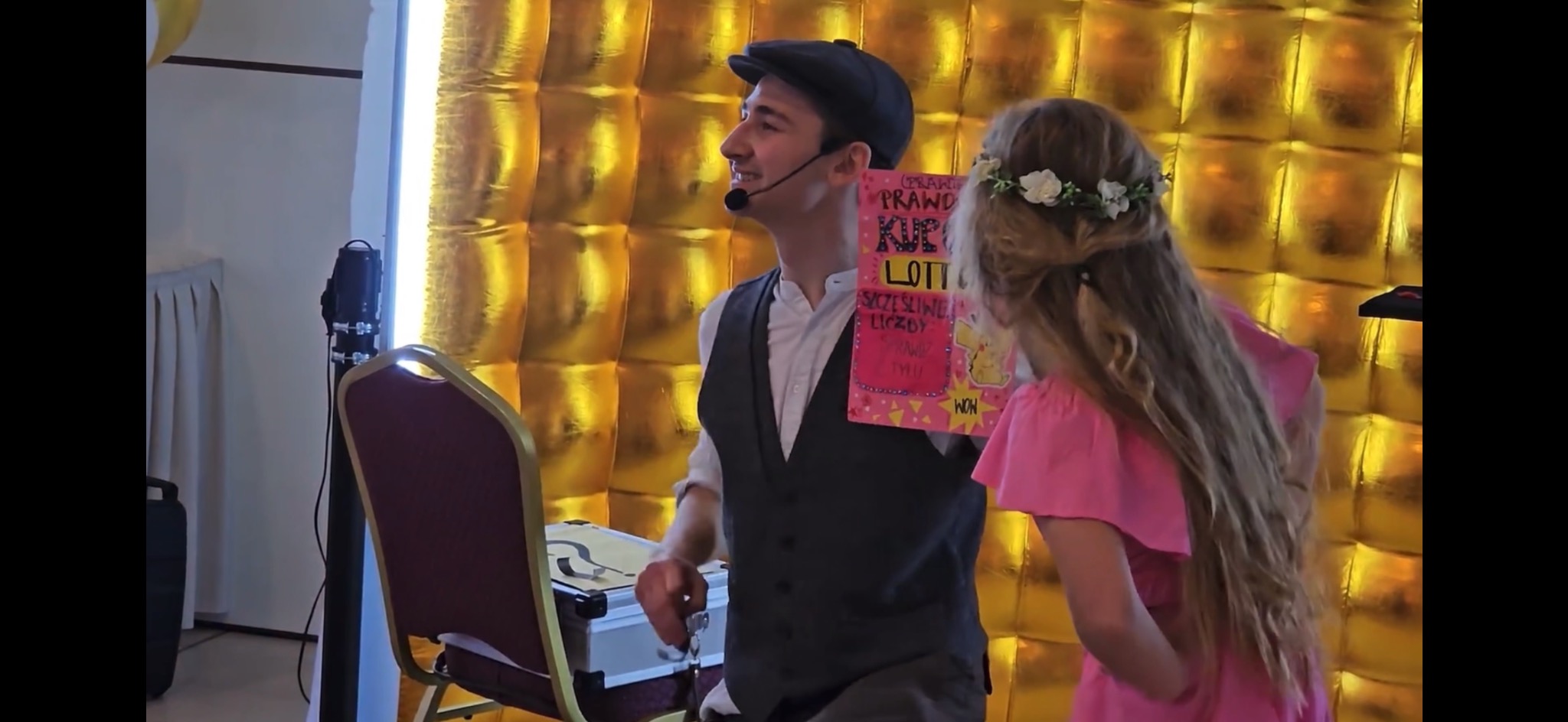 Uśmiechnięty iluzjonista w kaszkiecie prezentuje dziewczynce w wianku na głowie rekwizyt z napisem 'LOTTO' na tle złotej ścianki.