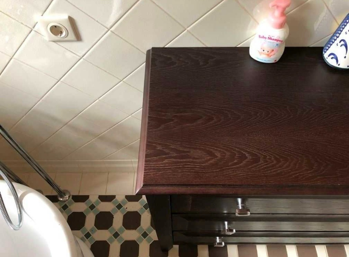 Widok z góry na fragment łazienki z ciemnobrązową komodą z trzema szufladami z kryształowymi uchwytami, białą wanną, biało-beżowymi płytkami na ścianach i wzorzystą podłogą z płytek.