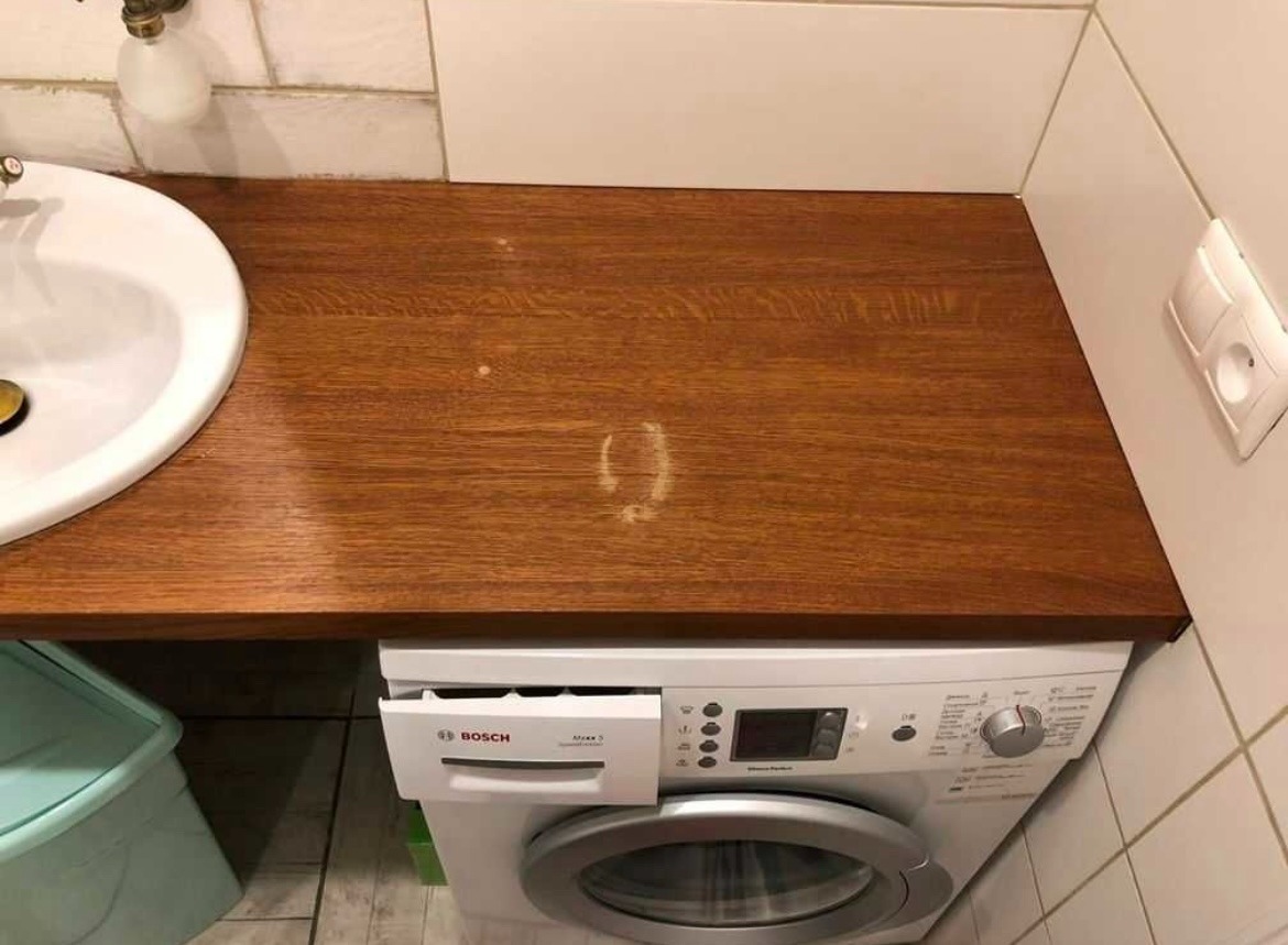 Blat drewniany nad pralką w łazience, obok umywalki, z widocznymi śladami użytkowania.