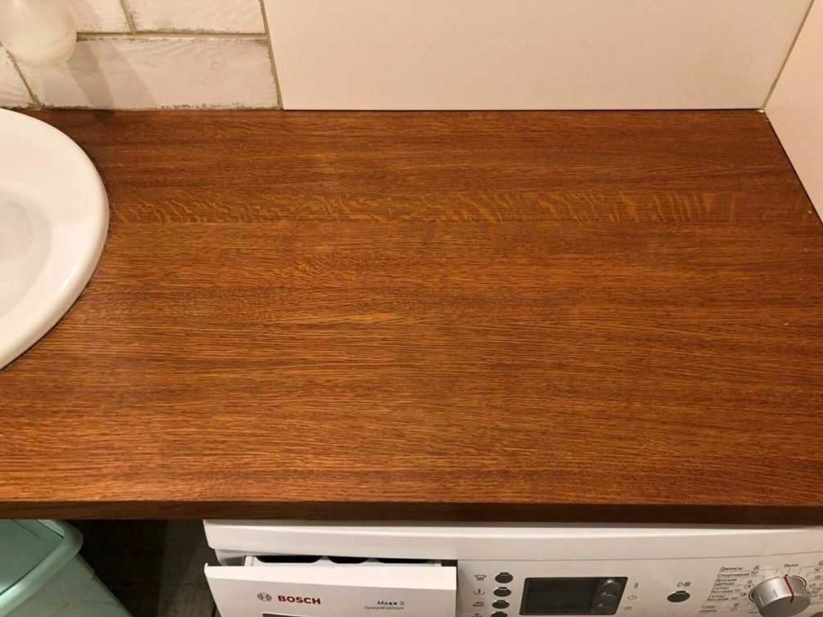 Blat z ciemnego drewna nad pralką w łazience, widoczna umywalka i białe płytki na ścianie.