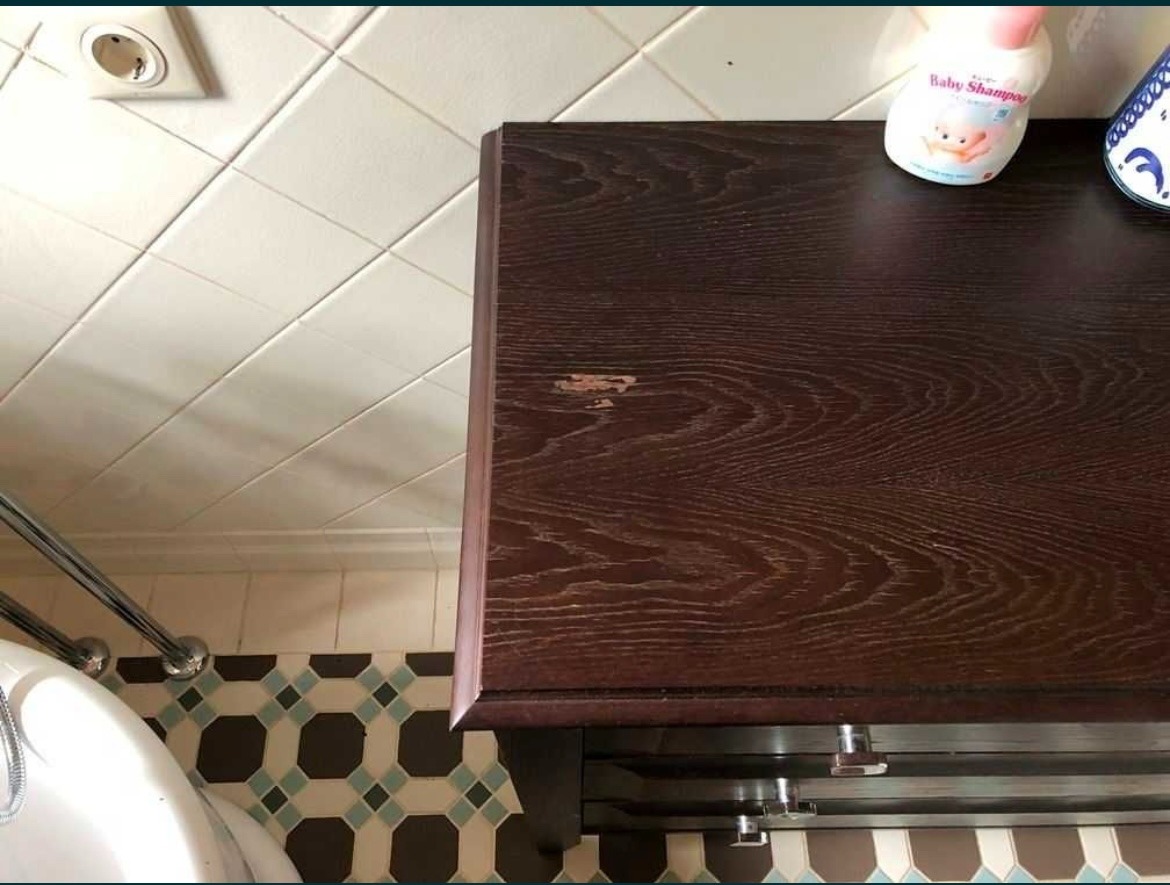 Ciemnobrązowa drewniana szafka łazienkowa z widocznym ubytkiem na blacie, obok biała butelka szamponu dla dzieci, na tle białych płytek i wzorzystej podłogi.