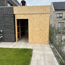 Magic Corp. - Konstrukcja z płyt OSB z otworem drzwiowym, prawdopodobnie garaż lub budynek gospodarczy, w trakcie budowy, widoczny szkielet drewniany i osoba na dachu, na tle zabudowy jednorodzinnej.