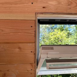 Magic Corp. - Fragment drewnianej ściany z zamontowanym oknem dachowym marki Velux, widok z zewnątrz, jasne drewno, widoczne łączenia desek i detale montażu.