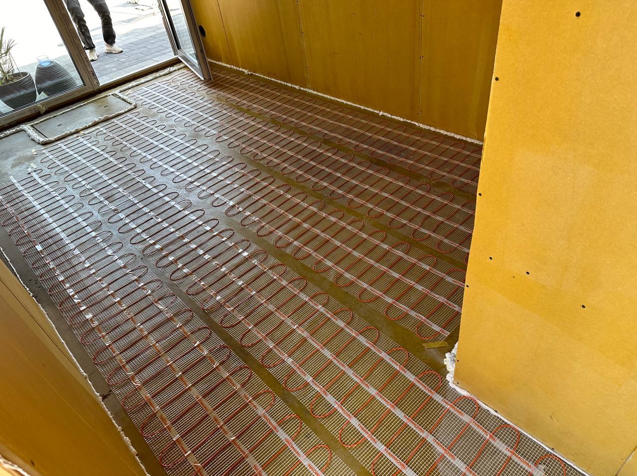 Rozłożona mata grzewcza z czerwonymi przewodami na podłodze w pomieszczeniu z żółtymi ścianami wykończonymi płytami, widoczne przygotowania do wylewki.