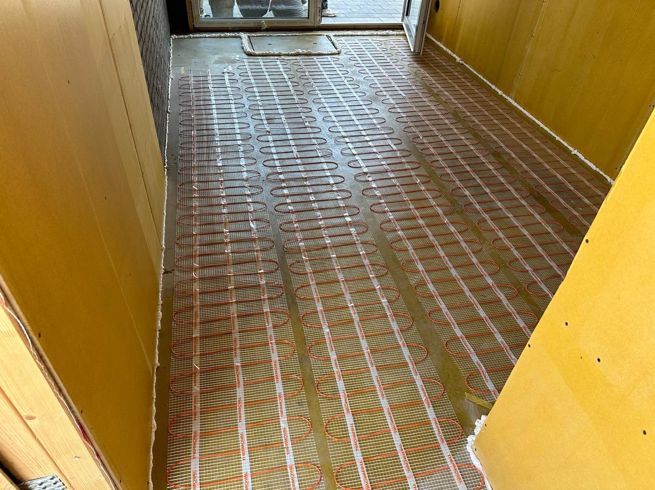 Rozłożona mata grzewcza z pomarańczowymi kablami na podłodze przygotowanej do wylewki, widoczne płyty kartonowo-gipsowe przy ścianach i fragment okna z doniczkami.