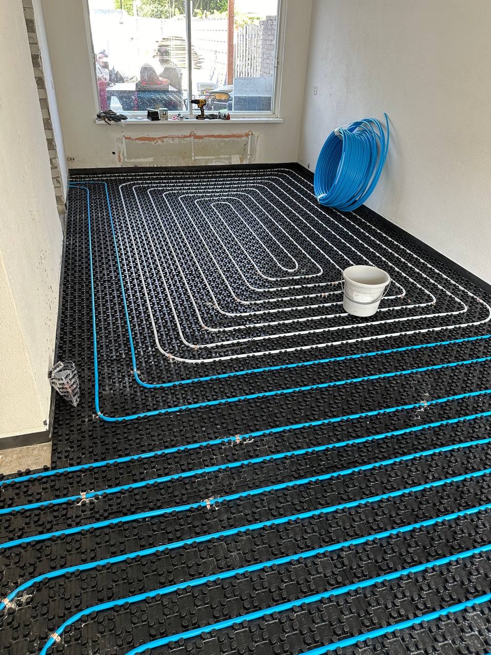 Instalacja ogrzewania podłogowego z białych i niebieskich rurek na czarnej macie izolacyjnej, widok z góry, w tle okno z narzędziami i wiadro.