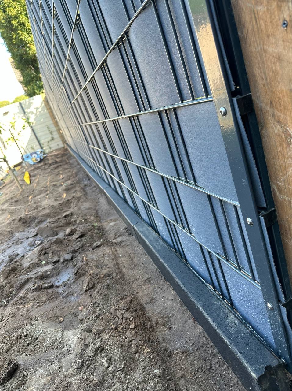 Fragment nowo postawionego, nowoczesnego ogrodzenia panelowego z szarym wypełnieniem, widoczne mocowania i podmurówka betonowa, teren wokół jeszcze niezagospodarowany z widoczną ziemią.