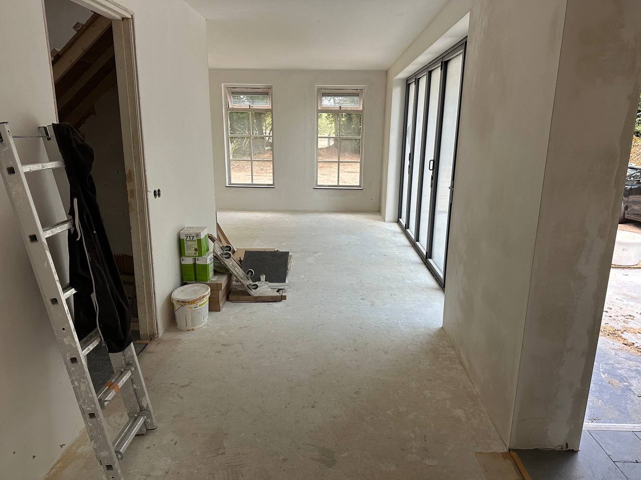 Wnętrze pokoju po szpachlowaniu ścian i podłogi, widoczna drabina, okna, drzwi przesuwne, wiadro z farbą i puszki z gładzią.