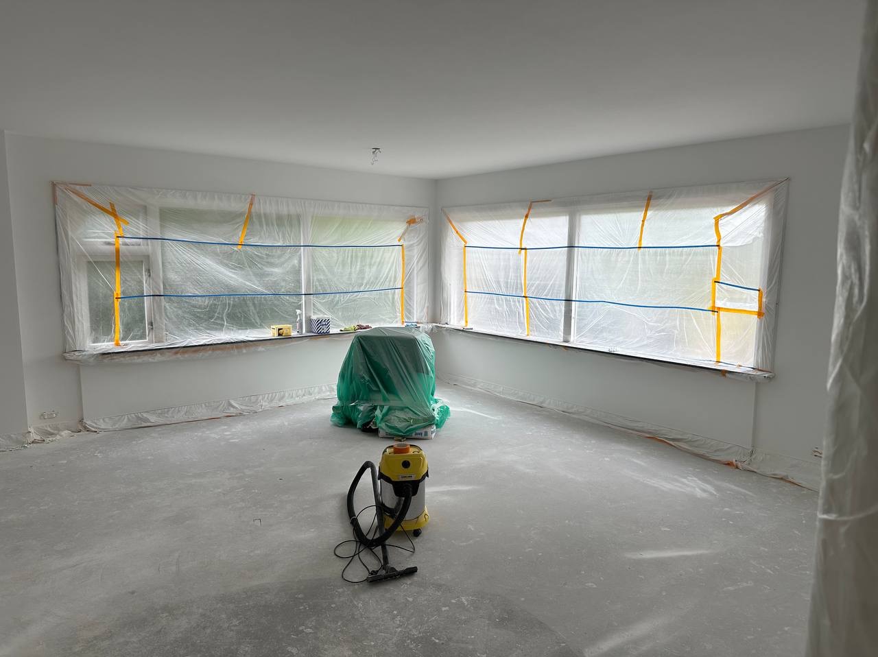 Pokój przygotowany do malowania: okna zabezpieczone folią malarską z taśmą, podłoga pokryta warstwą kurzu, odkurzacz przemysłowy w rogu, mebel zakryty zieloną folią ochronną.