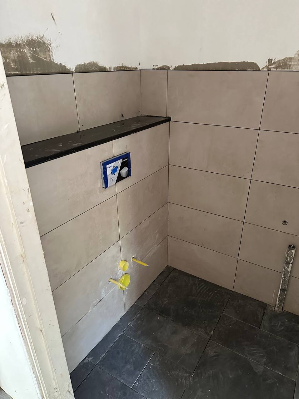 Wnętrze łazienki w trakcie wykańczania: ułożone jasne płytki ścienne, ciemna podłoga, widoczne elementy instalacji sanitarnej, w górnej części ściany pozostałości po pracach wykończeniowych.