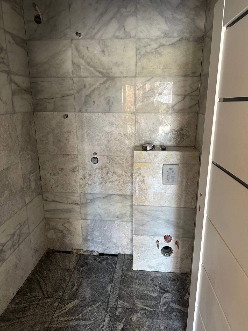 Wnętrze łazienki w trakcie remontu, widoczne marmurowe płytki na ścianach, ciemna podłoga z płytek, niezamaskowane otwory na armaturę i częściowo zamontowana instalacja podtynkowa.