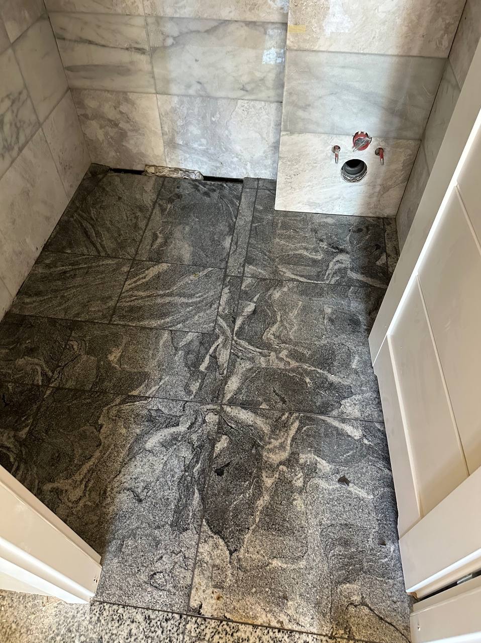 Podłoga łazienki w trakcie remontu z szarych płytek imitujących kamień, widoczne białe płytki na ścianie z wystającymi rurami i otworem odpływowym, dolna część białych drzwi po prawej stronie.