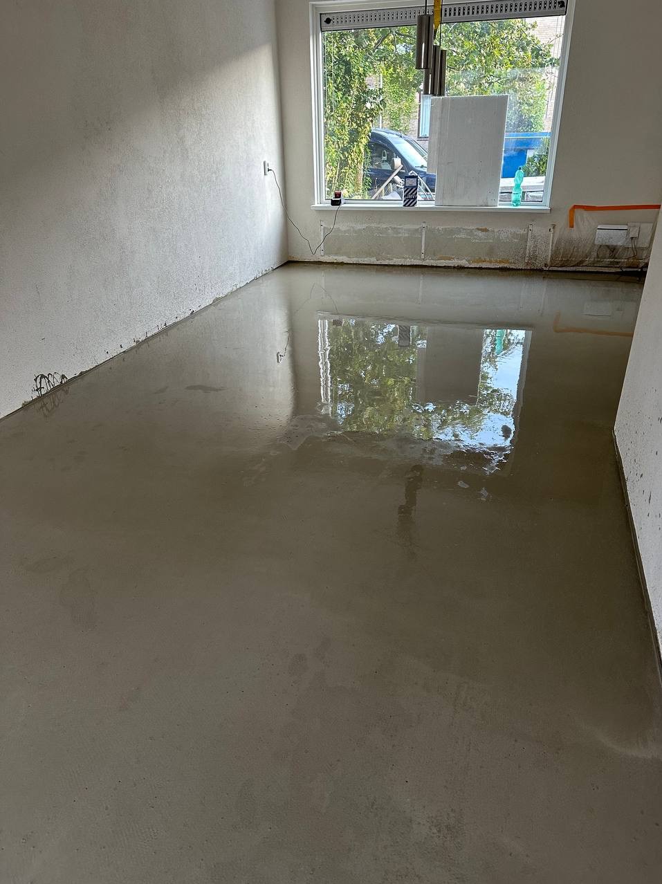 Wnętrze pokoju z wylewką samopoziomującą na podłodze, widoczne przygotowane ściany do dalszych prac wykończeniowych i okno z widokiem na zieleń.