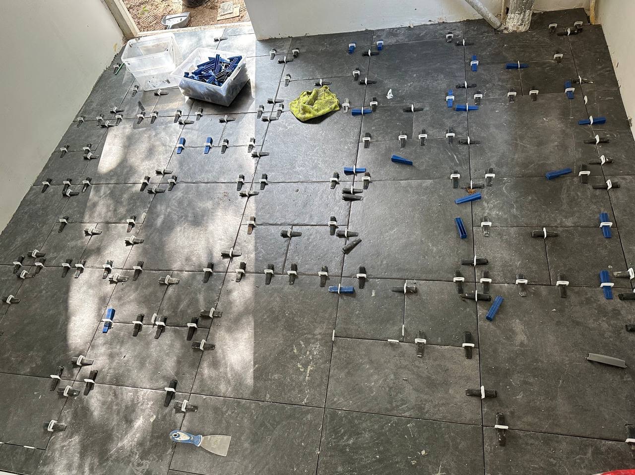 Układanie ciemnoszarych płytek podłogowych z systemem poziomowania: niebieskie kliny i białe krzyżyki dystansowe, widoczna szpachelka do rozprowadzania kleju i pojemnik z zapasowymi klinami, etap...