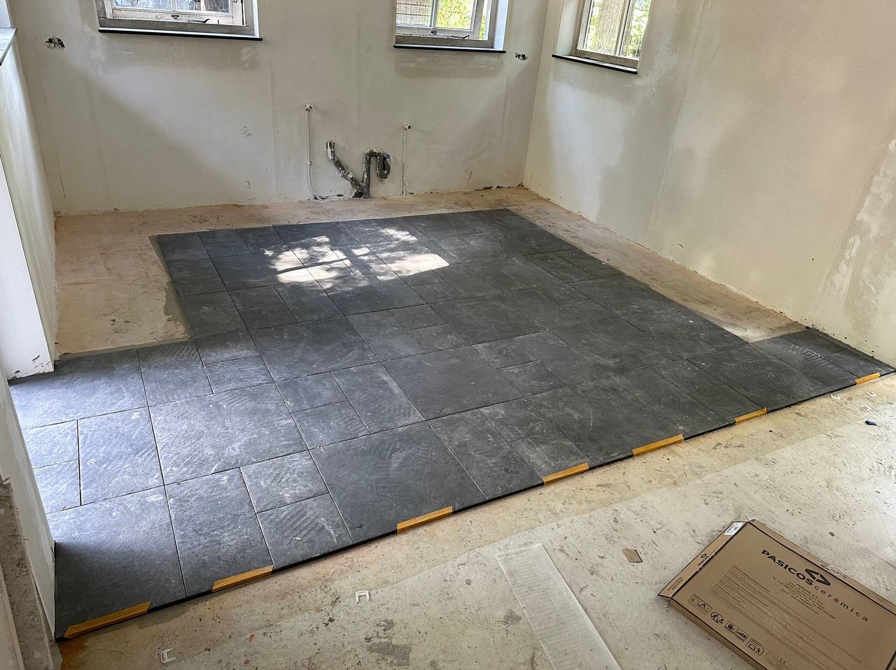 Układanie ciemnych, kwadratowych płytek podłogowych w surowym wnętrzu z widocznymi instalacjami, z użyciem żółtych klinów dystansowych przy ścianie, pudełko po płytkach Pasicos Ceramica w rogu.