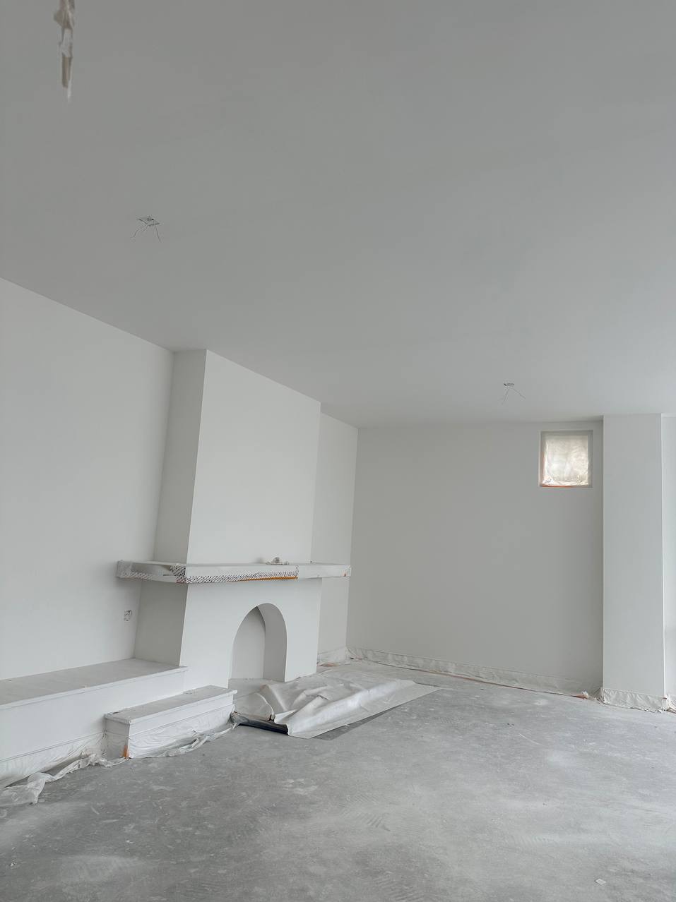 Białe wnętrze w trakcie prac wykończeniowych: ściany i kominek pomalowane na biało, podłoga zabezpieczona folią malarską, widoczne ślady szpachlowania i malowania.