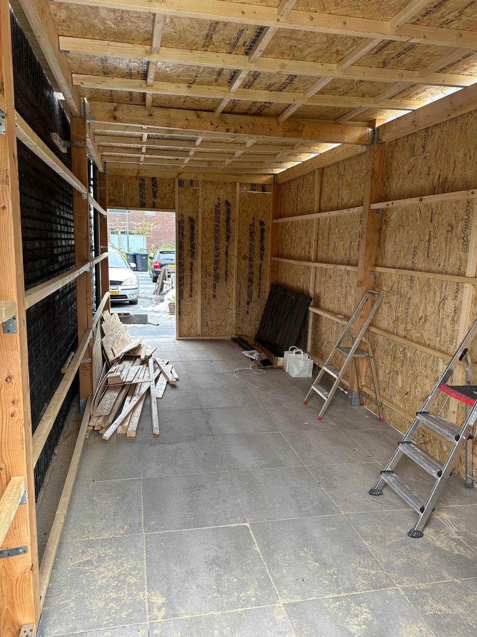 Wnętrze w trakcie budowy lub remontu, widoczne drewniane elementy konstrukcyjne ścian i sufitu, podłoga wyłożona płytkami, dwie drabiny, resztki drewna i narzędzia.