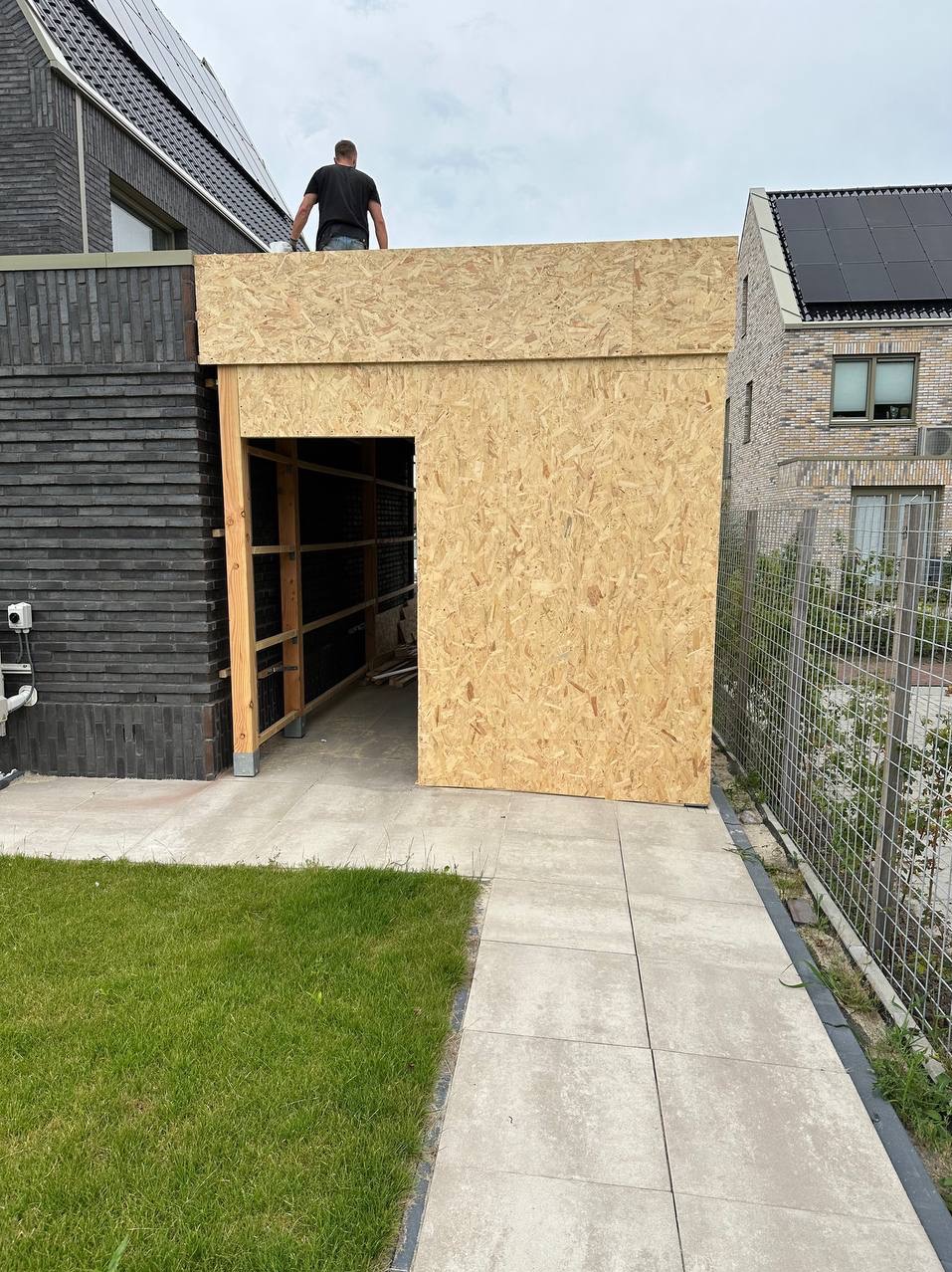 Konstrukcja z płyt OSB z otworem drzwiowym, prawdopodobnie garaż lub budynek gospodarczy, w trakcie budowy, widoczny szkielet drewniany i osoba na dachu, na tle zabudowy jednorodzinnej.
