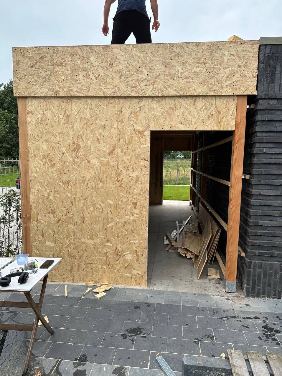Konstrukcja w trakcie budowy: ściana z płyty OSB z otworem drzwiowym, widoczne elementy drewniane, fragment muru z czarnej cegły, narzędzia na stole i osoba na dachu.