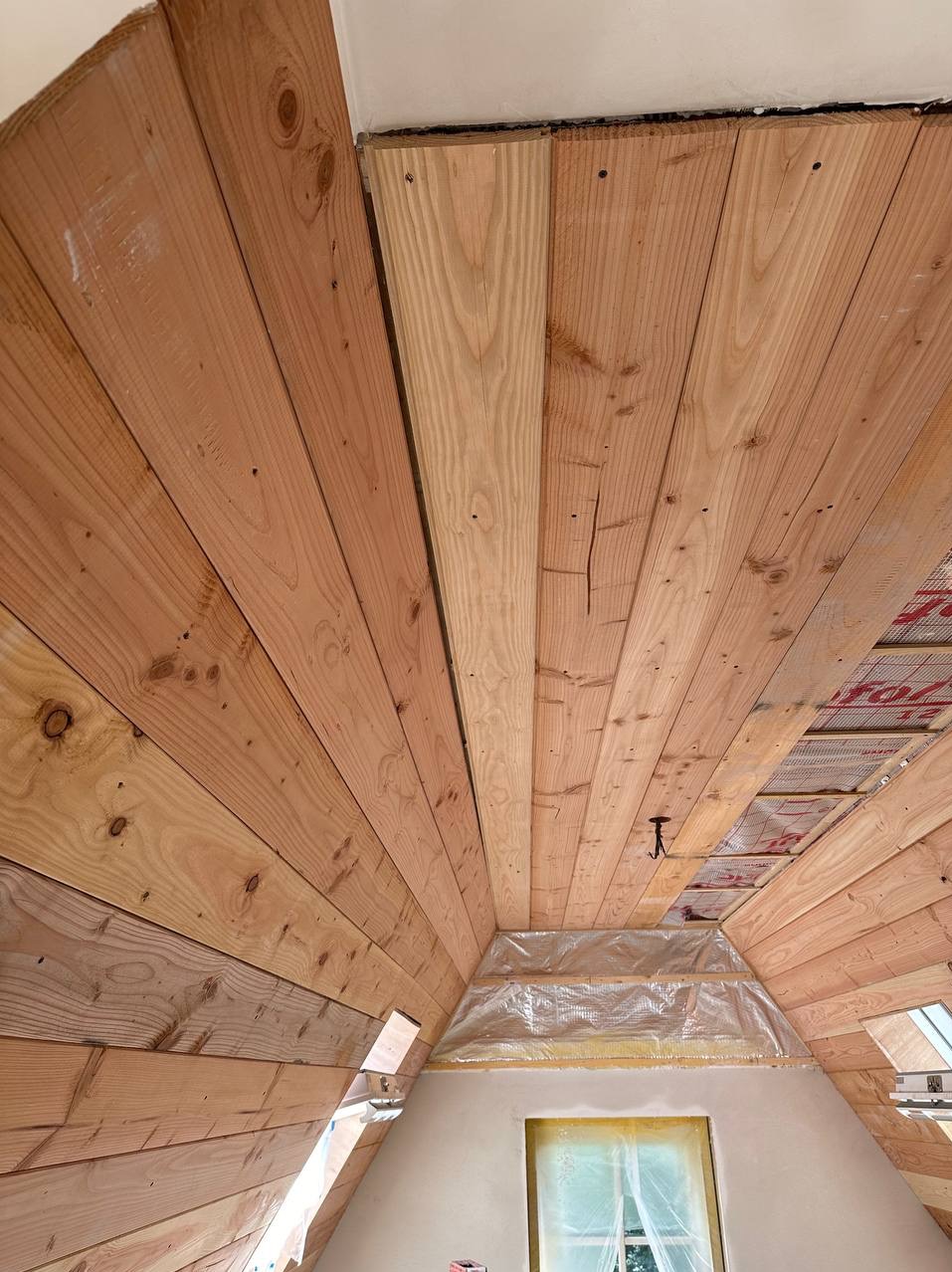 Drewniana boazeria na poddaszu w trakcie montażu, widoczne deski z naturalnego drewna, folia ochronna na ścianie i oknie, oraz fragment izolacji dachu.