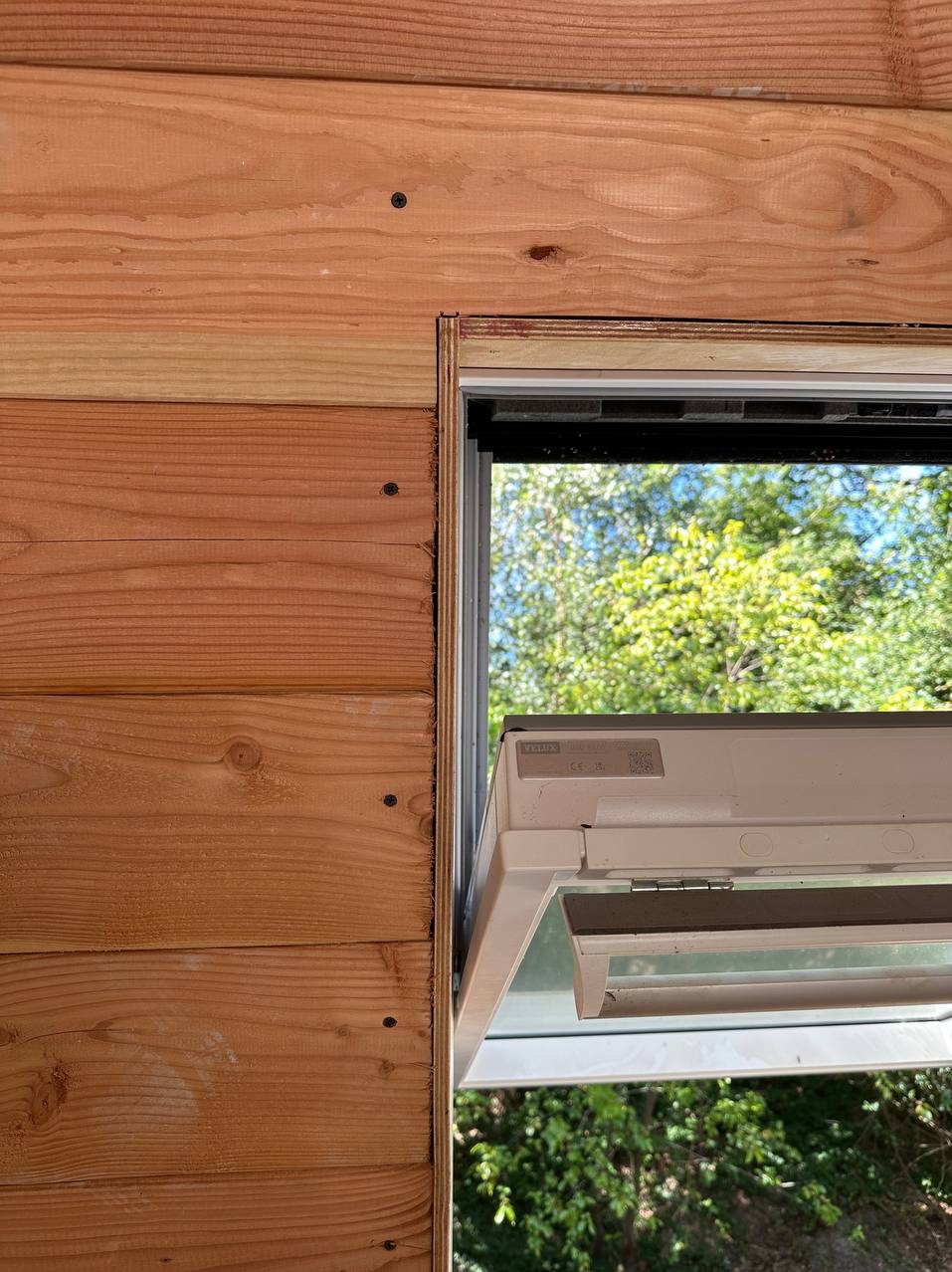 Fragment drewnianej ściany z zamontowanym oknem dachowym marki Velux, widok z zewnątrz, jasne drewno, widoczne łączenia desek i detale montażu.