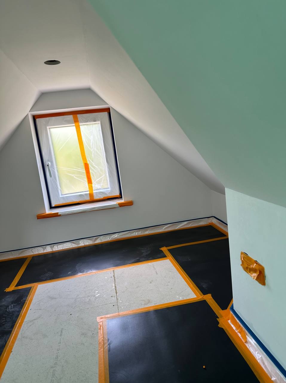 Pokój na poddaszu w trakcie remontu, okno zabezpieczone folią i taśmą, podłoga i ściany zabezpieczone folią malarską i taśmą malarską, widoczny surowy beton podłogi.