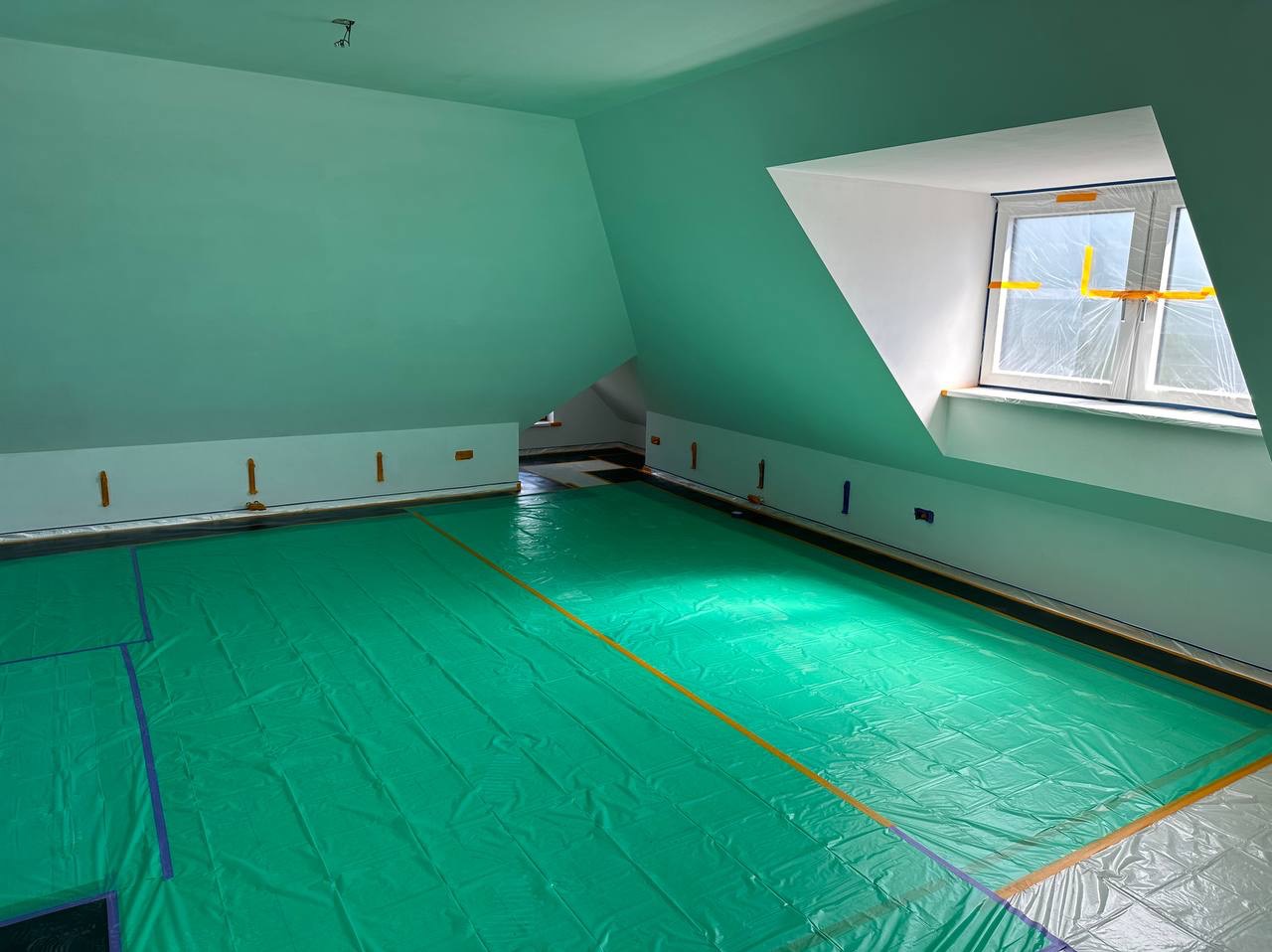 Pokój na poddaszu zabezpieczony folią malarską przed malowaniem ścian na kolor miętowy, okno zabezpieczone folią i taśmą. Widoczne ślady po gniazdkach elektrycznych.