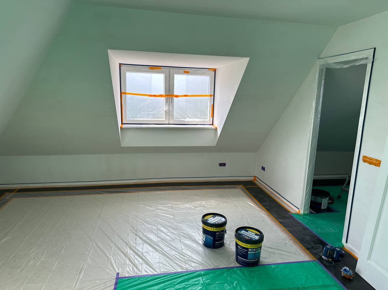 Pokój na poddaszu zabezpieczony folią i taśmą malarską przed malowaniem, widoczne dwa wiadra z farbą, ściany pomalowane na jasnozielony kolor, okno dachowe zabezpieczone folią.