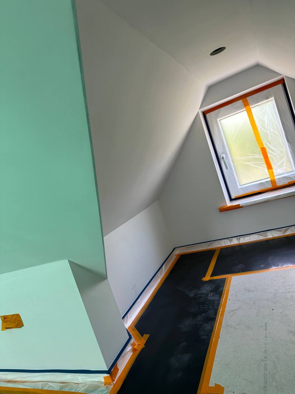 Pokój na poddaszu w trakcie zabezpieczania przed malowaniem: okno zaklejone folią, podłoga i krawędzie ścian zabezpieczone taśmą i folią malarską, część ściany pomalowana na zielono, reszta na biało.